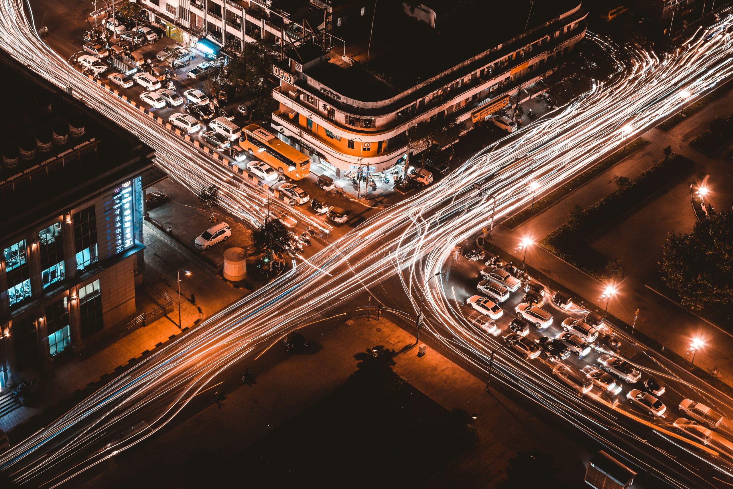 Aerial view of a busy city intersection at night with long light trails from moving vehicles. Surrounding buildings are illuminated, and parked cars line the streets.